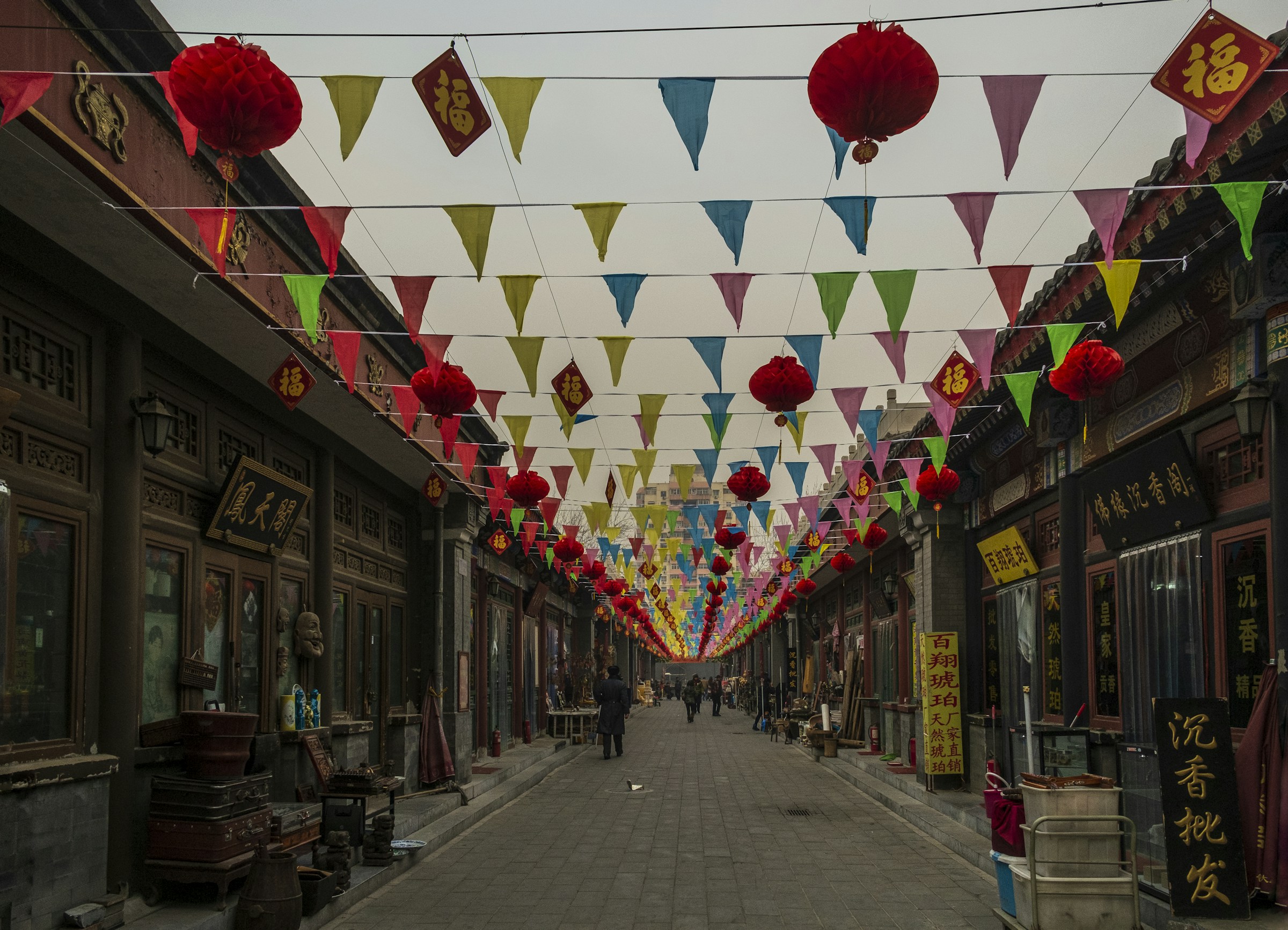 People walking through a Beijing market lane beneath lanterns and colored pennants